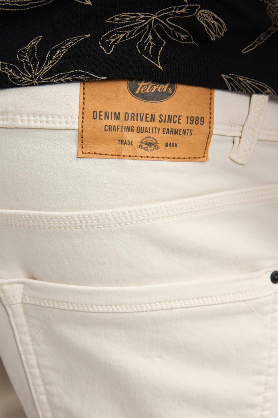 Ecru denim short van Petrol Industries®, met bruin leren merklabel en een zwart shirt met bladpatroon.