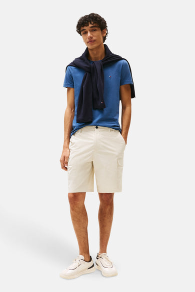 T-shirt bleu à manches courtes de Tommy Hilfiger, avec un pull sur les épaules, associé à des short beige et des baskets blanches.