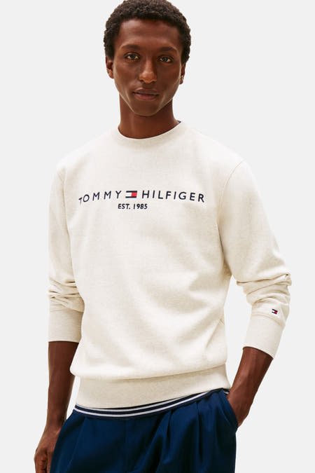 Gilet beige de TOMMY HILFIGER avec logo de marque sur la poitrine, associé à un pantalon bleu et des baskets blanches.