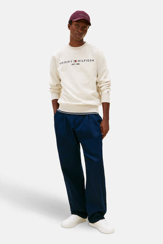 Beige sweater van TOMMY HILFIGER met merklogo op de borst, gecombineerd met blauwe pantalon en witte sneakers.
