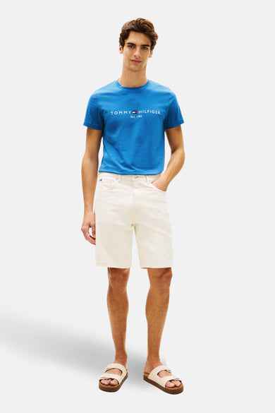 T-shirt bleu à manches courtes de Tommy Hilfiger, avec logo de la marque sur la poitrine, porté avec des shortis blancs et des sandales.