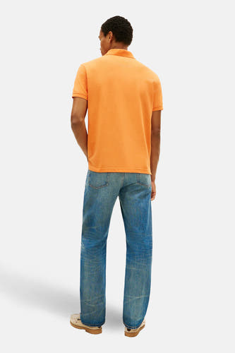 Oranje polo met korte mouwen van Tommy Hilfiger, gezien van achteren, met blauwe jeans en crèmekleurige schoenen.