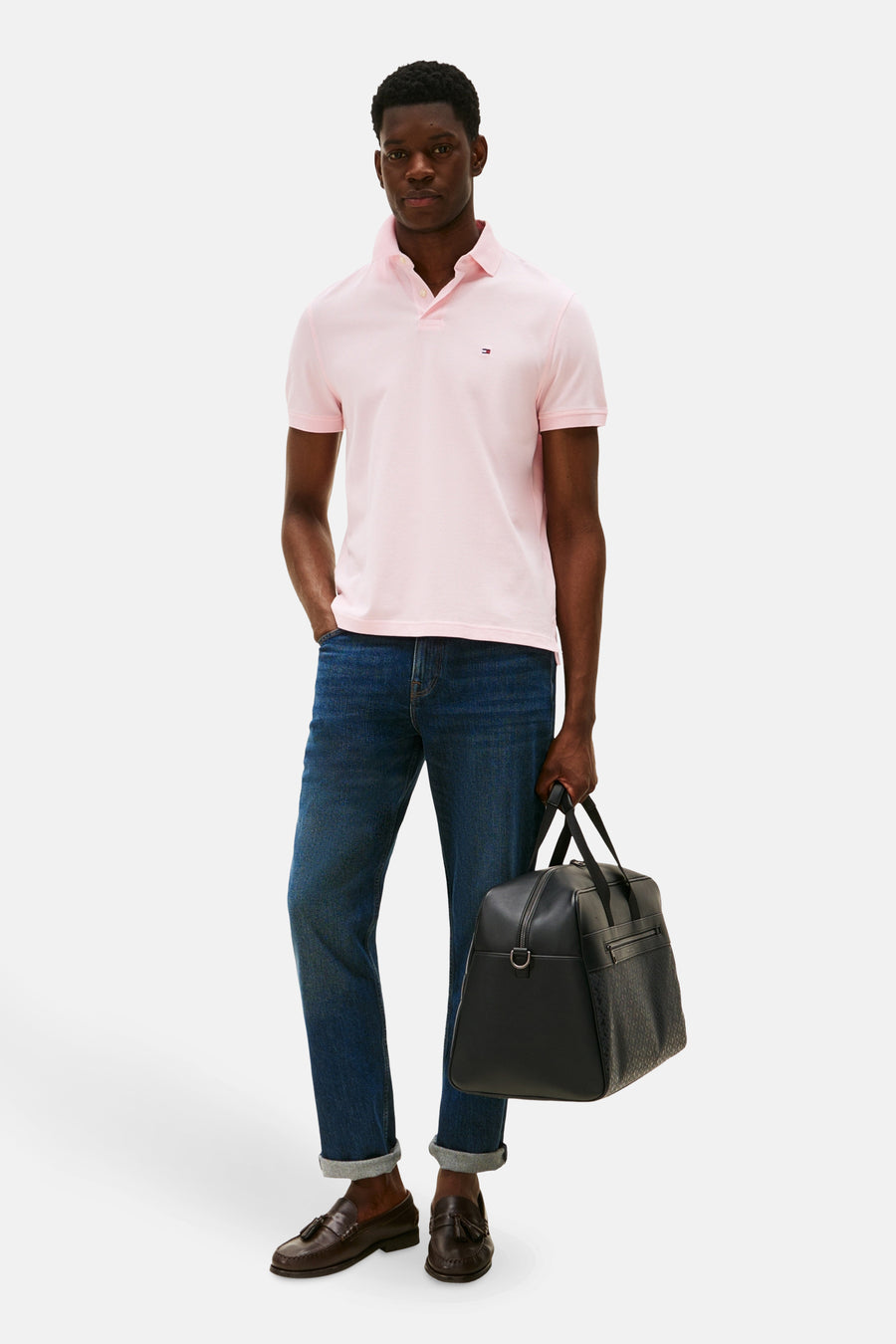 Roze polo met korte mouwen van TOMMY HILFIGER, gecombineerd met donkere jeans, leren loafers en een zwarte reistas.
