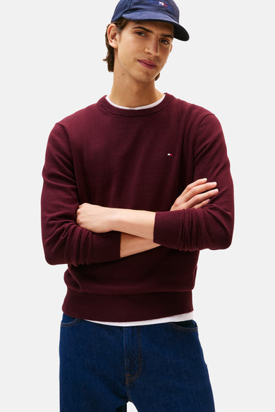 Bordeauxkleurige trui met ronde hals van TOMMY HILFIGER, met gekruiste armen en donkerblauwe jeans.