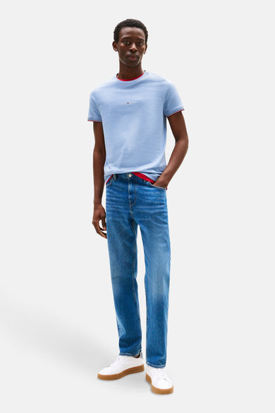 T-shirt bleu à manches courtes de TOMMY HILFIGER, avec un bord rouge et associé à des jeans et des chaussures blanches.