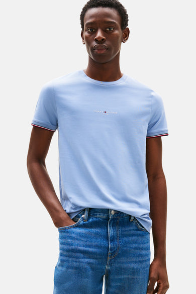 T-shirt bleu à manches courtes de TOMMY HILFIGER, avec un bord rayé rouge et blanc sur les manches, porté avec des jeans bleus.