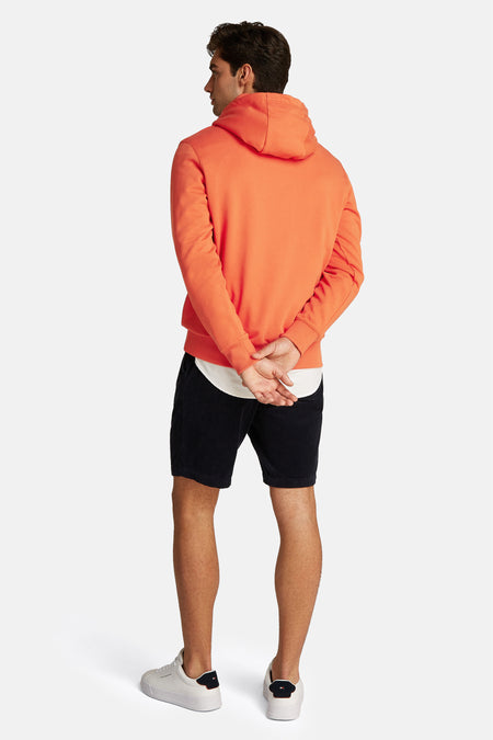 Pull-over orange Tommy Hilfiger avec capuche, côté dos, associé à des shorts de corduroy bleu marine et des baskets blanches.
