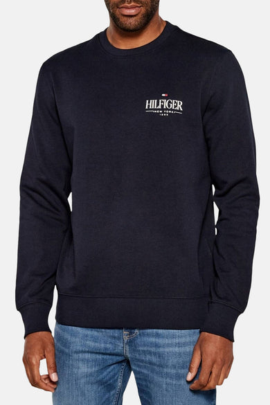 Blauwe sweater van TOMMY HILFIGER met wit logo op de borst en lange mouwen, gedragen met een jeans.
