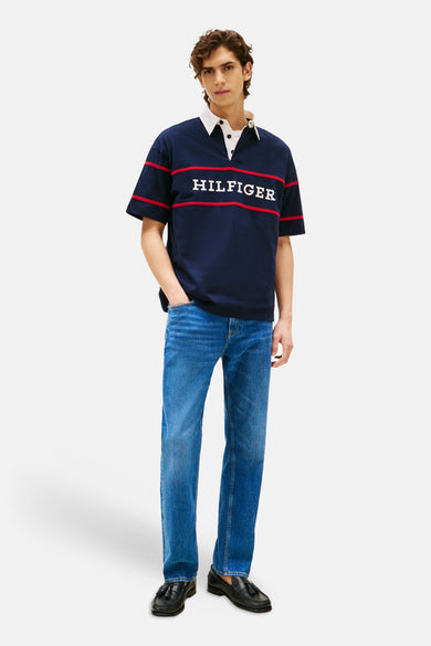 Blauw poloshirt met korte mouwen van Tommy Hilfiger, met rode strepen en 'Hilfiger' in witte letters, gedragen met blauwe jeans en zwarte loafers.