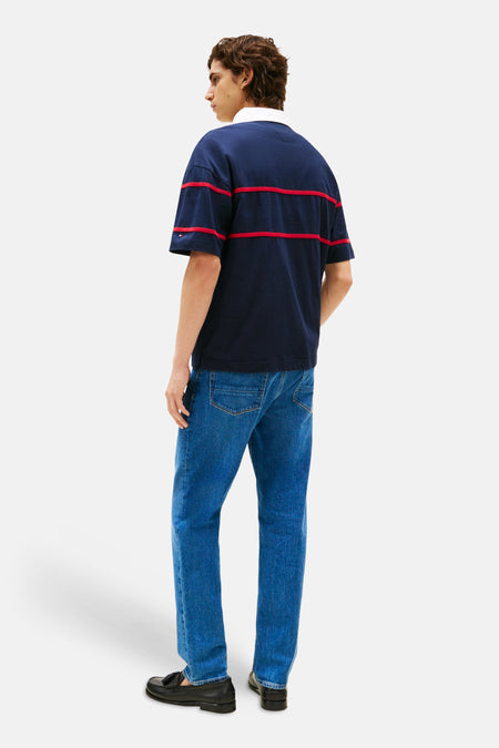 Blauwe polo met korte mouwen van Tommy Hilfiger, met rode strepen en gedragen met blauwe jeans en zwarte loafers.
