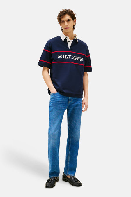 Blauwe polo met korte mouwen van Tommy Hilfiger, met witte kraag en rode strepen en de tekst 'HILFIGER'. Gedragen met blauwe jeans en zwarte schoenen.