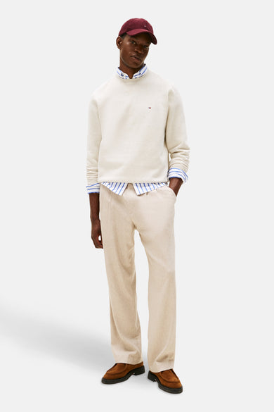 Ecrukleurige sweater met ronde hals van TOMMY HILFIGER, gedragen over een gestreept overhemd, met beige broek en bruine schoenen.
