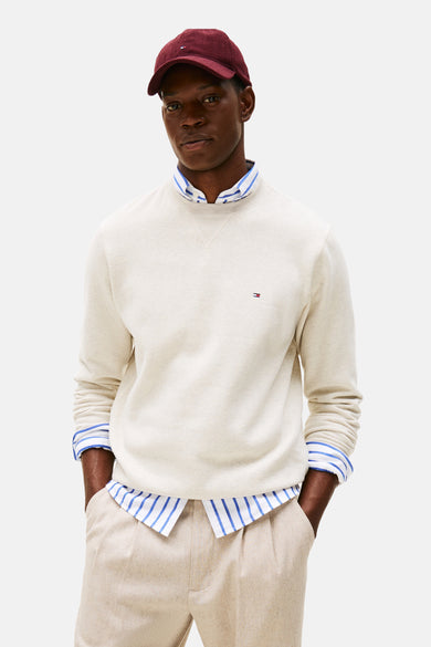 Ecru sweater met ronde hals van TOMMY HILFIGER, gedragen over een gestreept overhemd, met een bordeauxrode pet.
