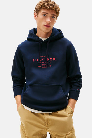 Blauwe sweater met kap van Tommy Hilfiger, met een rood logo op de borst, gedragen met een kaki broek.
