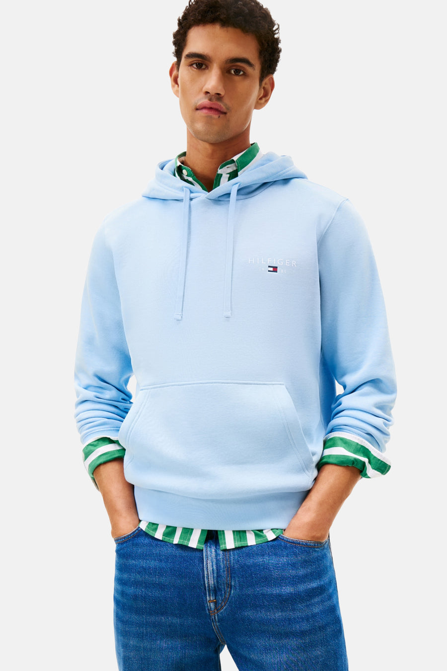 Blauwe sweater met kap van Tommy Hilfiger, met trekkoorden en buidelzak, gedragen over een gestreept overhemd, met jeans.
