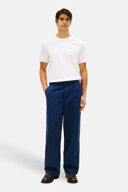 Wit T-shirt met korte mouwen van TOMMY HILFIGER met lichtblauw logo op de borst, gedragen door een man met donkerblauwe broek en bruine loafers.
