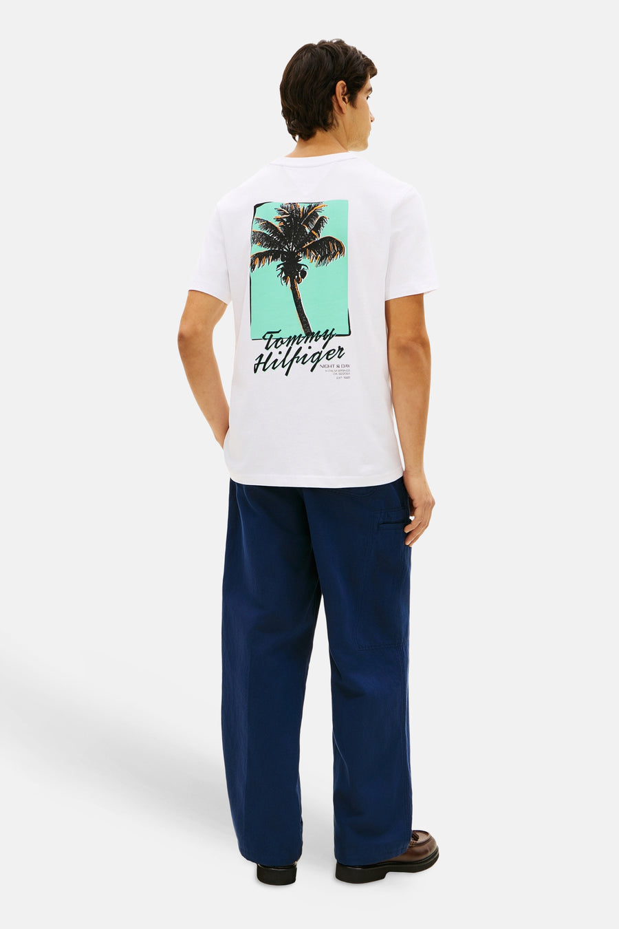 T-shirt blanc à manches courtes TOMMY HILFIGER avec grand imprimé palmier et logo dans le dos, porté par un homme en pantalon bleu foncé.