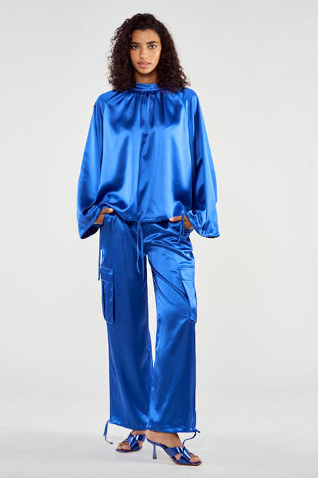 Blauwe cargobroek van NOOR BY NORA GHARIB, met zakken en trekkoordjes aan de pijpen, gedragen met blauwe hakken.
