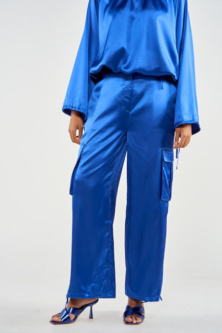 Blauwe cargobroek van NOOR BY NORA GHARIB, met satijnen textuur en wijde pijpen, gecombineerd met blauwe top en hakken.
