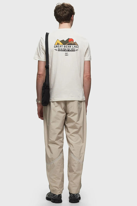 Ecru T-shirt van Napapijri, met bergen en logo op de rug, gecombineerd met beige broek en zwarte tas.
