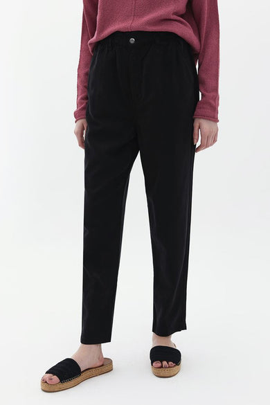Pantalon noir d'OXXO, porté avec un gilet rose et des sandales tressées.