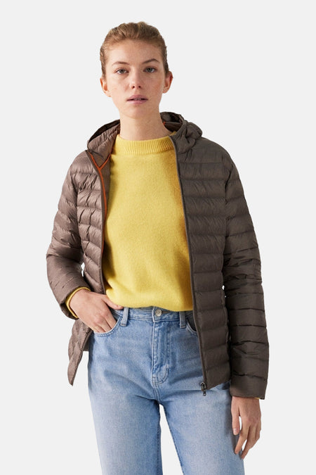 Vrouw in bruine jas met gele trui en jeans tegen witte achtergrond.