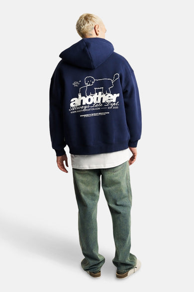 Pull-over bleu avec capuche d'ANOTHER COTTON LAB, avec une impression blanche sur l'arrière, associé à un T-shirt blanc et des jeans verts.