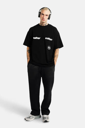 T-shirt noir à manches courtes d'ANOTHER COTTON LAB, avec une impression de texte blanche et porté avec un pantalon noir et des baskets grises.