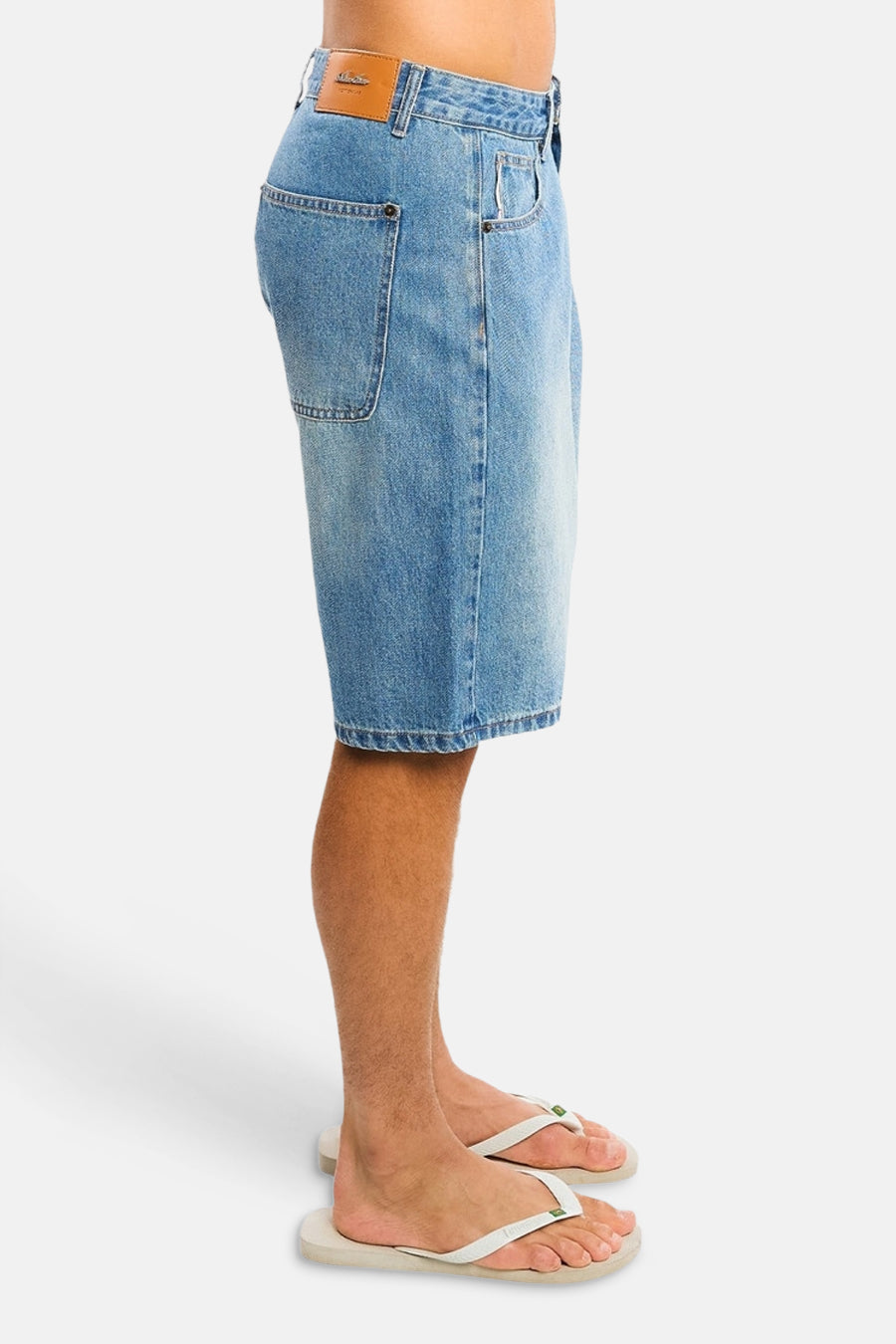 Lichtblauwe denim jeansshort van ANOTHER COTTON LAB, zijprofiel, met vervaagde was, achterzakken en bruin leerlabel, gedragen met witte slippers.