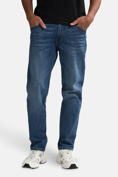 Taps toelopende jeans van middelblauw denim van Petrol Industries, met twee handen in de zakken en witte sneakers.
