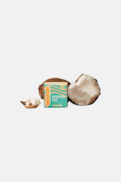 Shampoo bar in een turquoise doos met beige golfpatroon van WONDR CARE, tussen halve kokosnoten en een houten schaaltje met kokosstukjes.