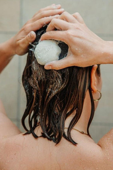 Witte vaste douchegel van WONDR CARE schuimt op nat donker haar van een persoon in de douche, met een gouden oorring.