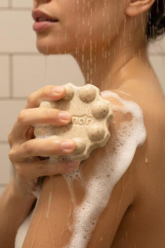 Een lichtbeige douchegelreep met scrubdeeltjes van WONDR CARE, rond met nopjes en tekst 'ndr', schuimend op een arm onder de douche.