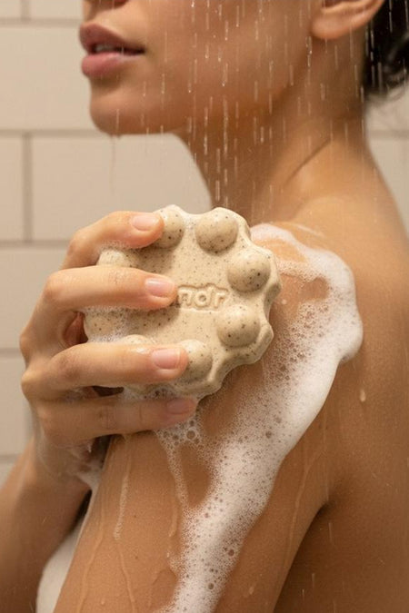 Een lichtbeige douchegelreep met scrubdeeltjes van WONDR CARE, rond met nopjes en tekst 'ndr', schuimend op een arm onder de douche.