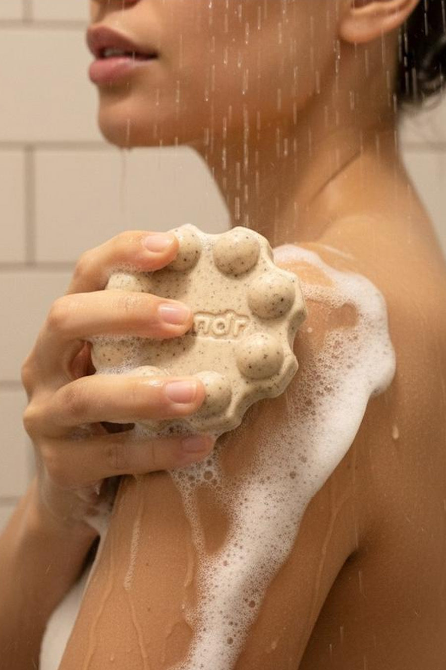 Een lichtbeige douchegelreep met scrubdeeltjes van WONDR CARE, rond met nopjes en tekst 'ndr', schuimend op een arm onder de douche.