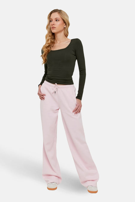 Vrouw met blond haar in groene top en roze wijde joggingbroek, staand.
