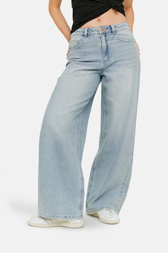 Lichtblauwe wijde jeans, wit sportief schoeisel, model staat met handen in zakken.