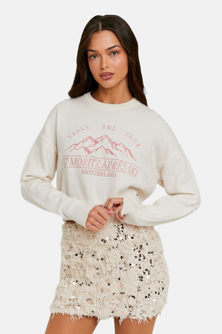 Pull-over blanc avec col rond de TALLY WEIJL, imprimé d'un club de ski rose et porté avec une jupe brillante.