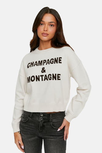 Ecrukleurige trui met ronde hals van TALLY WEIJL, met opschrift 'Champagne & Montagne', gecombineerd met een grijze jeans.
