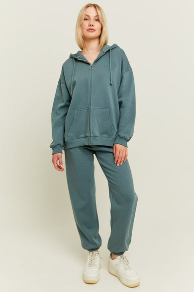 Gilet turquoise de TALLY WEIJL avec capuchon, fermeture éclair et poches à l'avant, porté avec un pantalon de jogging coordonné et des baskets blanches.