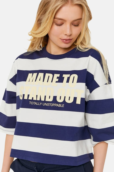 T-shirt bleu de TALLY WEIJL avec des rayures blanches horizontales et le texte jaune "MADE TO STAND OUT" et "TOTALLY UNSTOPPABLE", porté par une femme blonde.