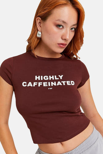 Bruin T-shirt met korte mouwen van TALLY WEIJL, met de tekst "HIGHLY CAFFEINATED" en gedragen met zilveren oorbellen.
