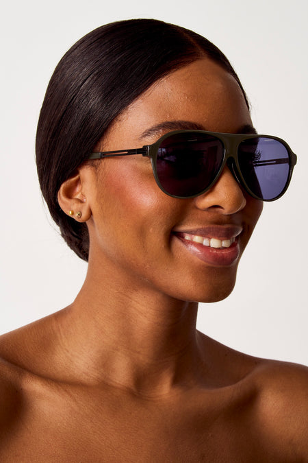 Lunettes de soleil vertes de SUNSET STUDIO, portées par une femme souriante avec des boucles d'oreilles dorées et une peau brune.