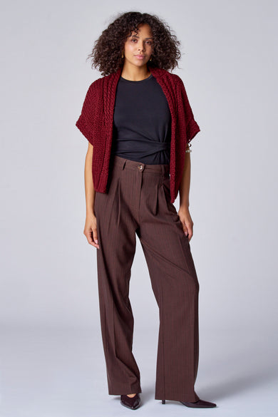 Vrouw in rode vest, zwarte top en bruine pantalon voor een grijze achtergrond.