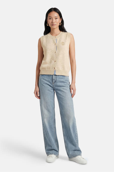 Pull beige ADORE, sans manches, avec fermeture à boutons et écusson, combiné avec un jean bleu et des baskets blanches.