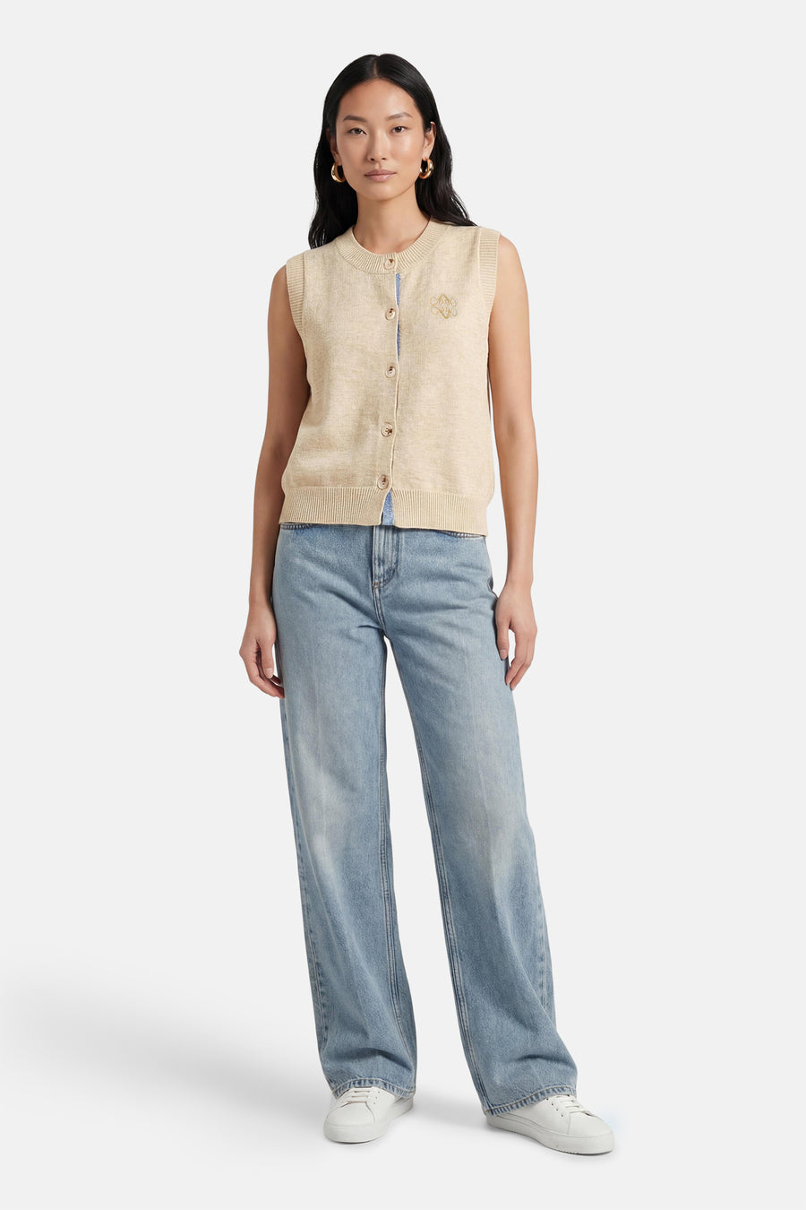 Beige trui van ADORE, mouwloos, met knoopsluiting en embleem, gecombineerd met blauwe jeans en witte sneakers.