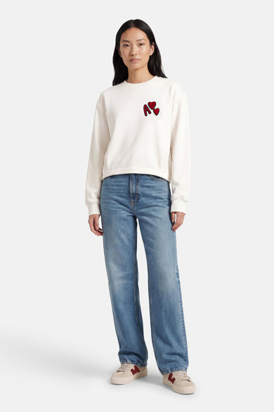 Witte sweater met ronde hals van ADORE, versierd met een rode letter en hartjes, gedragen met blauwe jeans en crèmekleurige sneakers.
