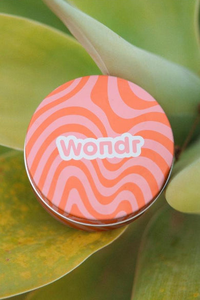 Multicolor verzorgingsproduct van WONDR CARE, met een golvend patroon, geplaatst op een groene plant.
