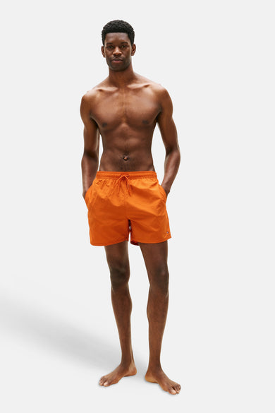 Oranje zwemshort van Tommy Hilfiger, gedragen door een man met zijn handen in de zakken, op een witte achtergrond.