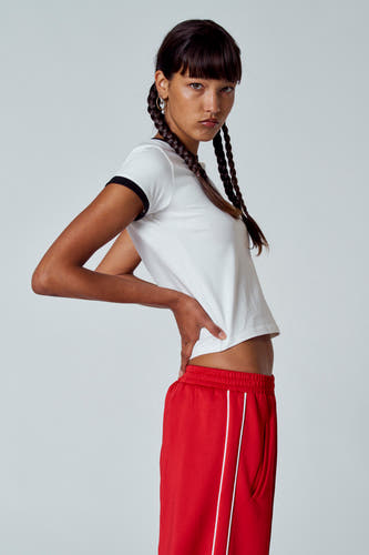 T-shirt blanc avec des manches courtes et des détails noirs d'Unwind, porté avec un pantalon de jogging rouge et une coiffure tressée.